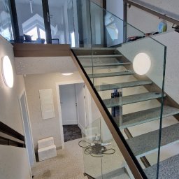 MGM STAL SPÓŁKA Z OGRANICZONĄ ODPOWIEDZIALNOŚCIĄ - Wewnętrzne schody z szarego kamienia z przezroczystą balustradą szklaną i metalową ramą, oświetlone okrągłymi lampami ściennymi, z widocznymi drzwiami i elementami wyposażenia wnętrza.