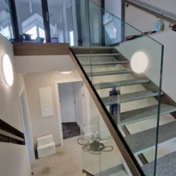 Wewnętrzne schody z szarego kamienia z przezroczystą balustradą szklaną i metalową ramą, oświetlone okrągłymi lampami ściennymi, z widocznymi drzwiami i elementami wyposażenia wnętrza.