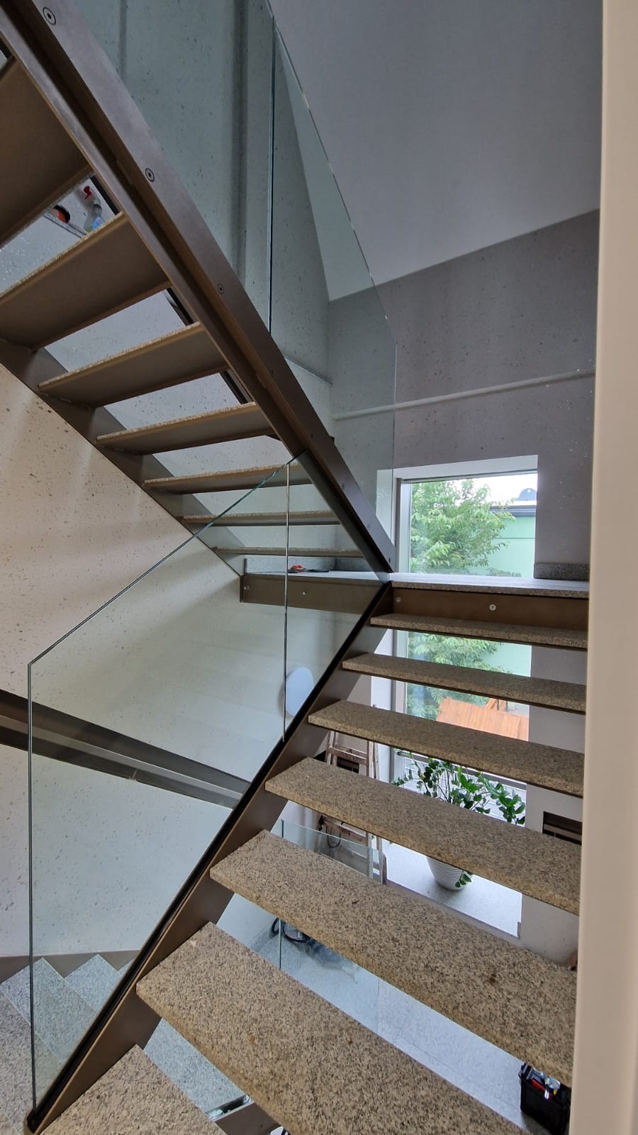 Nowoczesne schody z metalową konstrukcją i szklaną balustradą, widok z dołu, stopnie wykończone materiałem imitującym kamień, w tle okno z widokiem na zieleń.