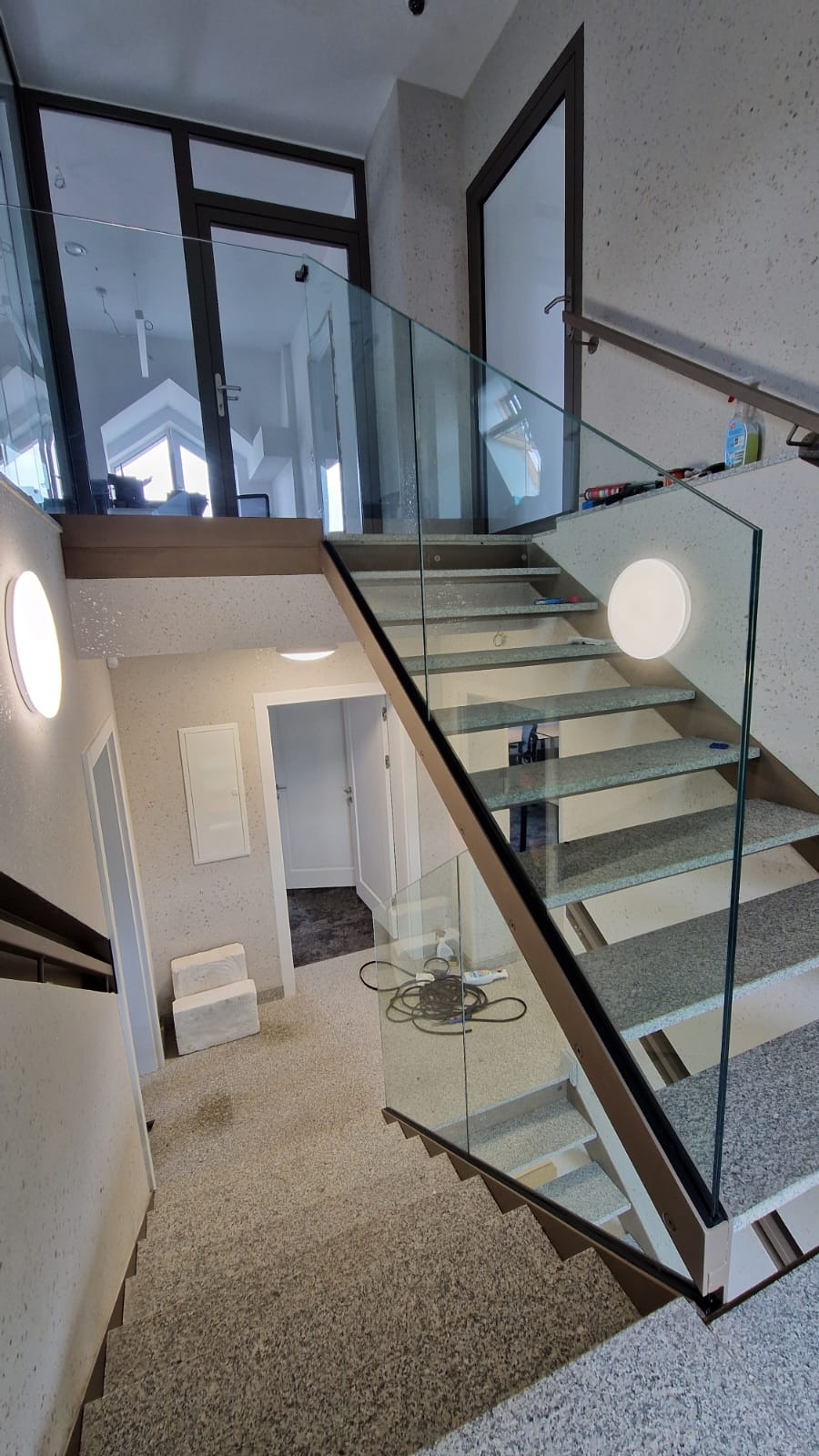 Wewnętrzne schody z szarego kamienia z przezroczystą balustradą szklaną i metalową ramą, oświetlone okrągłymi lampami ściennymi, z widocznymi drzwiami i elementami wyposażenia wnętrza.