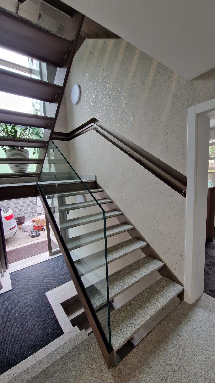 Wewnętrzne schody z metalową konstrukcją i szklaną balustradą w Pyskowicach, widoczne z dołu, z wejściem i roślinami w tle.