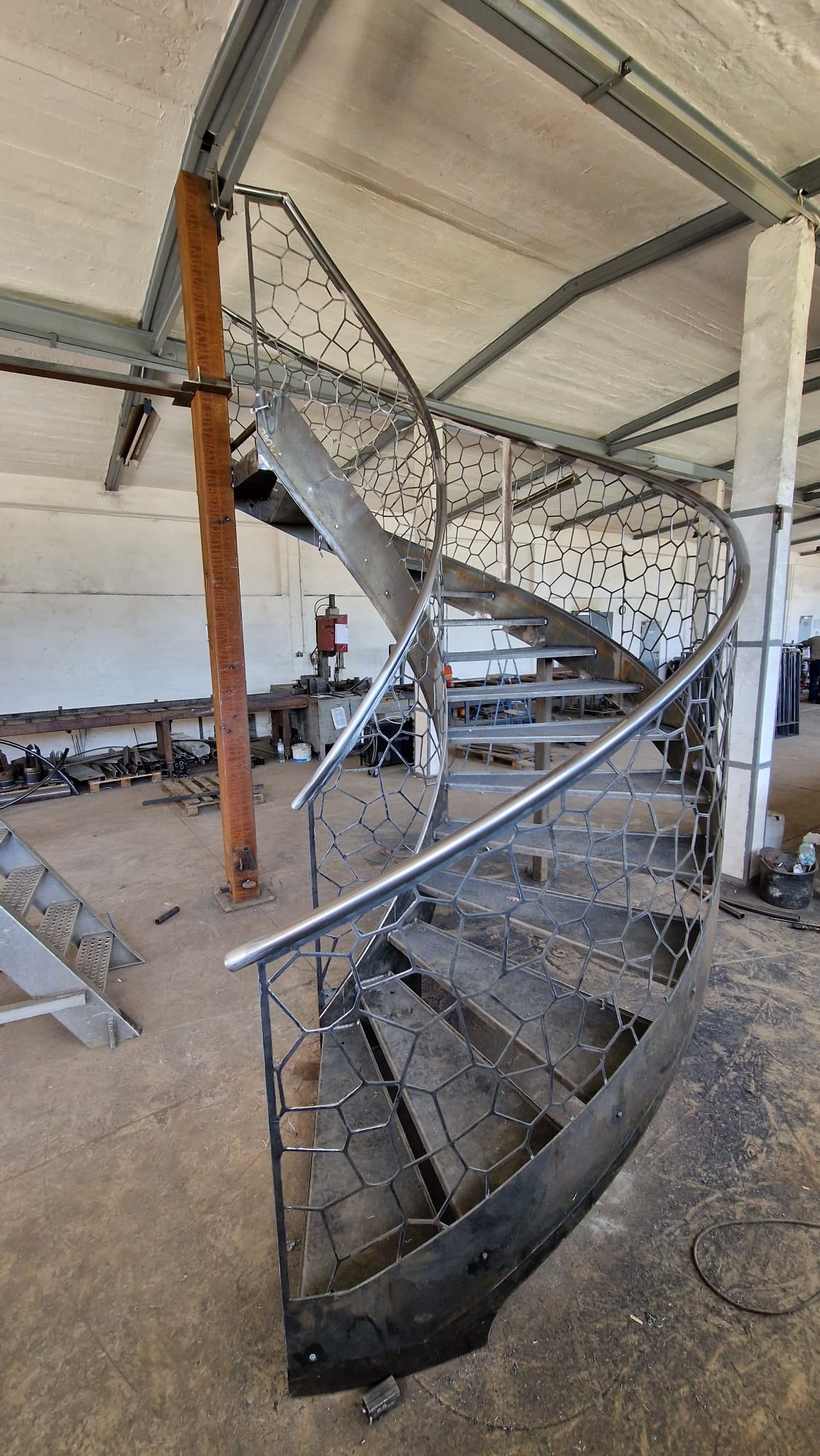 Metalowe schody spiralne z ażurową balustradą o wzorze plastra miodu, w trakcie montażu w hali warsztatowej, widoczne narzędzia i elementy konstrukcyjne w tle.