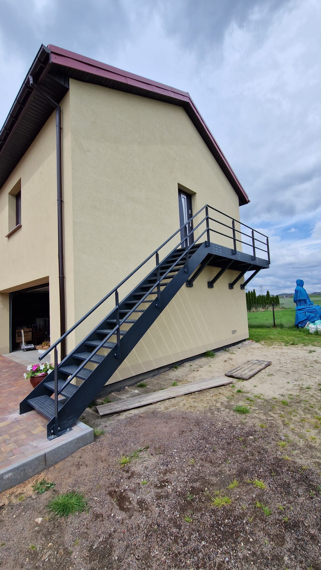 Zewnętrzne, czarne schody metalowe z balustradą prowadzące do drzwi na piętrze budynku o beżowej elewacji, z widocznym fragmentem dachu krytego czerwoną dachówką i otwartym garażem z boku.