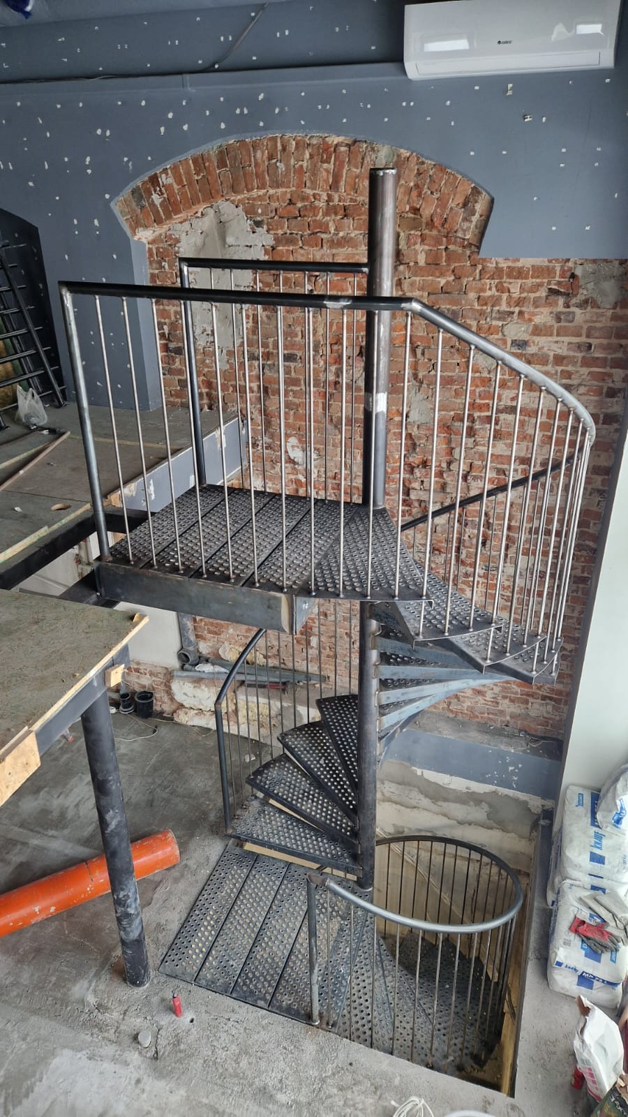 Metalowe schody spiralne z balustradą z prętów, w surowym stanie, na tle ceglanej ściany i szarego sufitu w trakcie remontu.