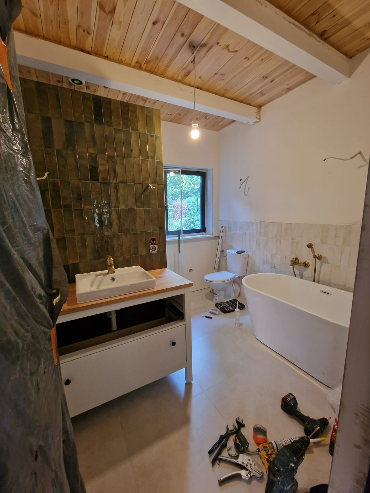 Remontowana łazienka z drewnianym sufitem, zielonymi płytkami na ścianie, białą umywalką na drewnianym blacie i narzędziami na podłodze.