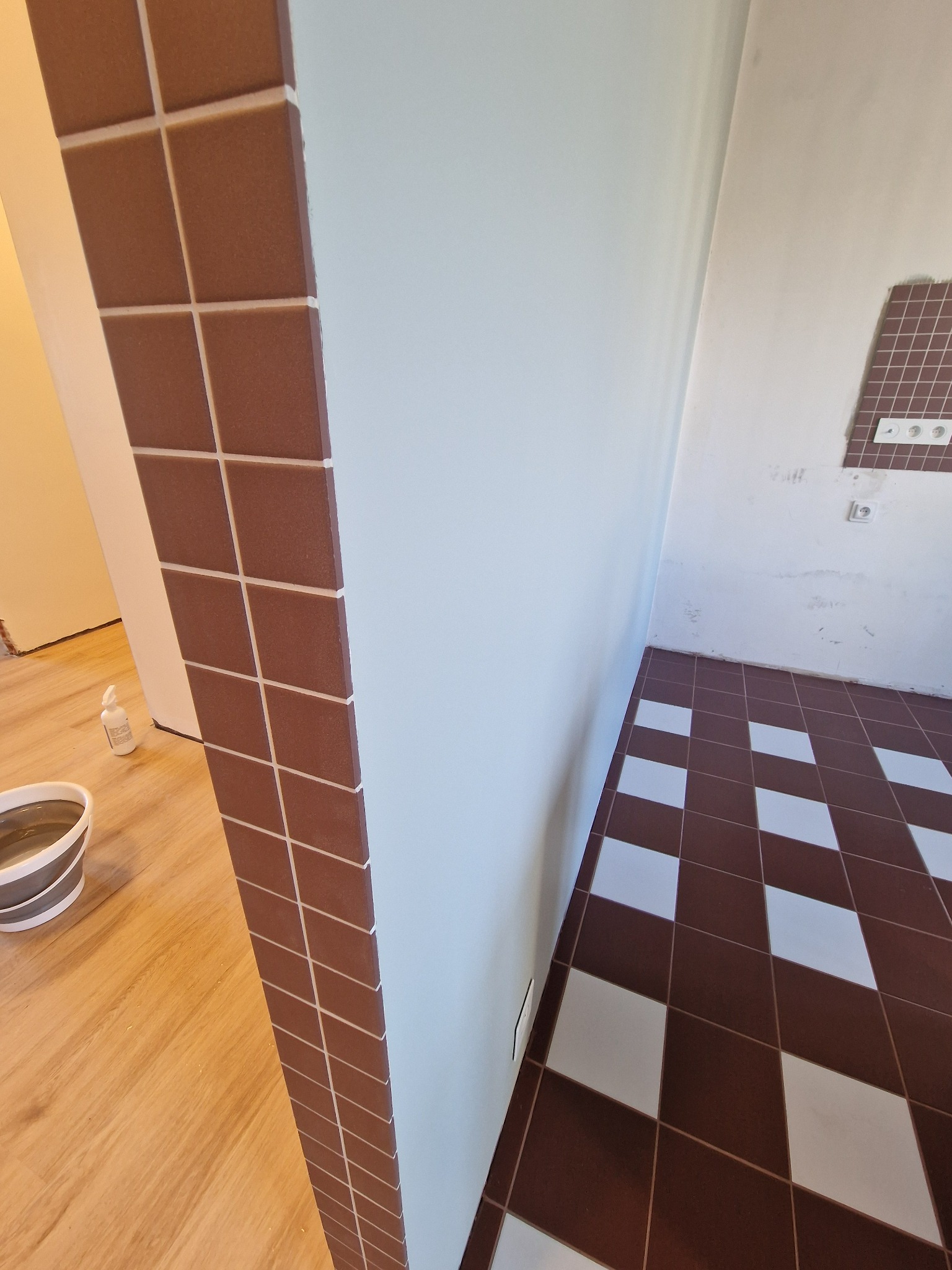 Brązowe płytki na ścianie i podłodze w romby, białe ściany, widoczny fragment podłogi drewnianej i wiadro. Częściowo wykończone gniazdka elektryczne.