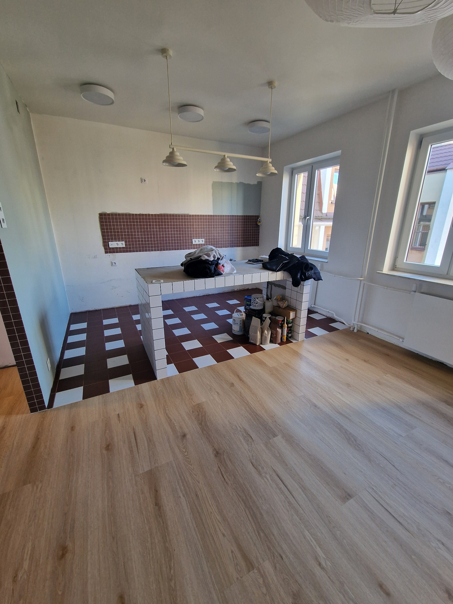 Wnętrze w trakcie prac wykończeniowych: ułożona podłoga, częściowo wykończona ściana z brązową mozaiką, wyspa wyłożona białymi płytkami. Widoczne narzędzia i materiały.