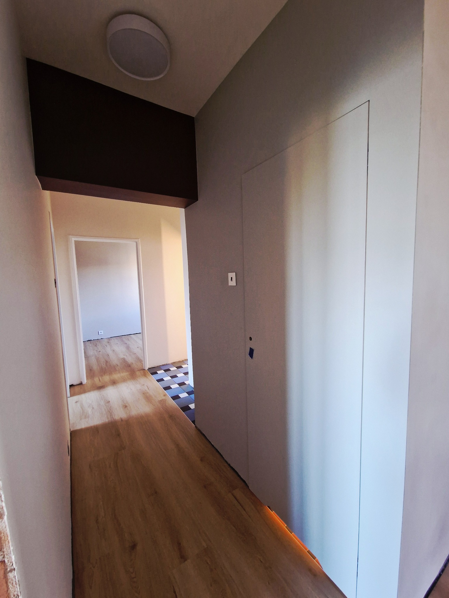 Korytarz po remoncie: panele podłogowe imitujące drewno, białe ściany, zabudowana szafa wnękowa. W tle widoczne przejście do innego pomieszczenia z szachownicą na podłodze.