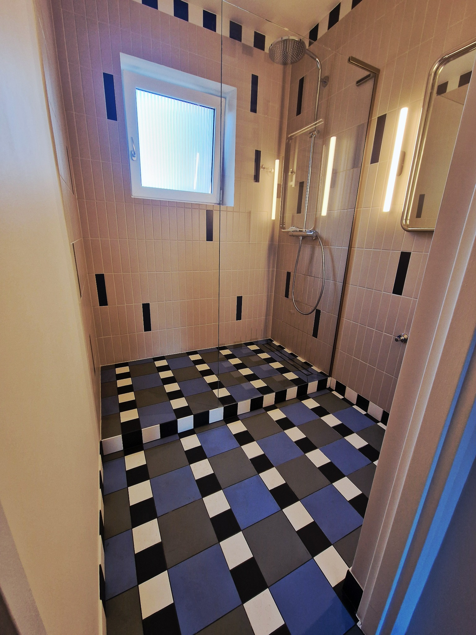 Nowoczesna łazienka z kabiną prysznicową, wykończona w beżowej i czarnej kolorystyce. Uwagę przykuwają wzorzyste, niebiesko-szaro-białe płytki podłogowe.