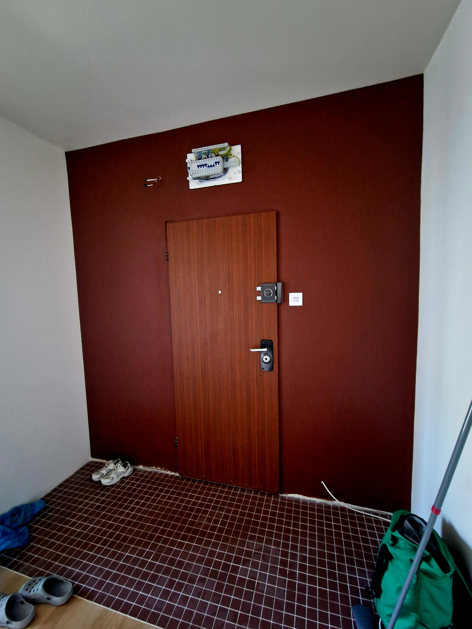 Korytarz po remoncie: brązowa ściana, drewniane drzwi z zamkiem, płytki podłogowe w kratkę. Widoczna skrzynka elektryczna i buty. Ukończone prace wykończeniowe.