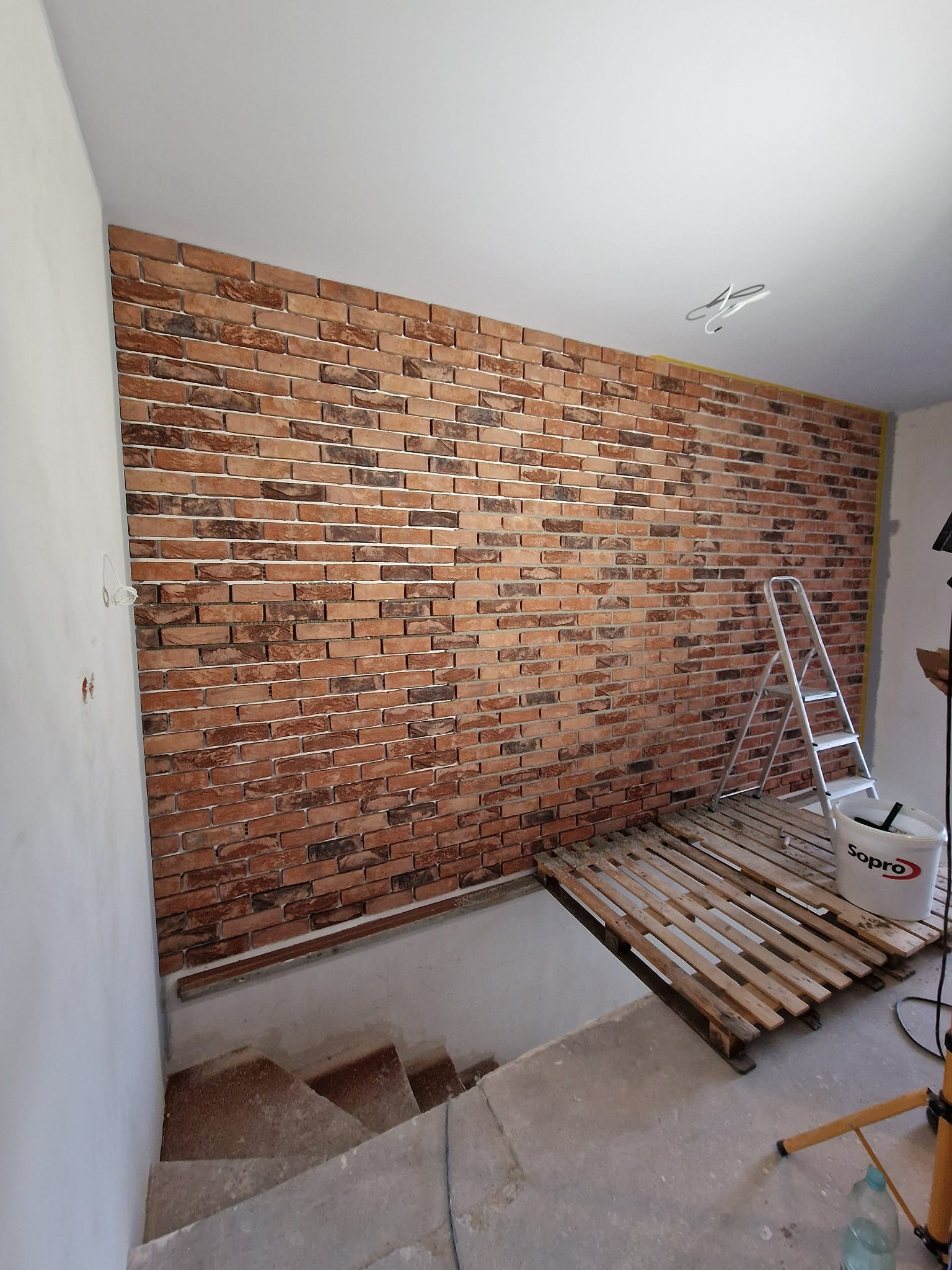 Ściana wykończona cegłą dekoracyjną w pomieszczeniu ze schodami, widoczny fragment drabiny i wiadro z zaprawą Sopro. Częściowo wykończone wnętrze.