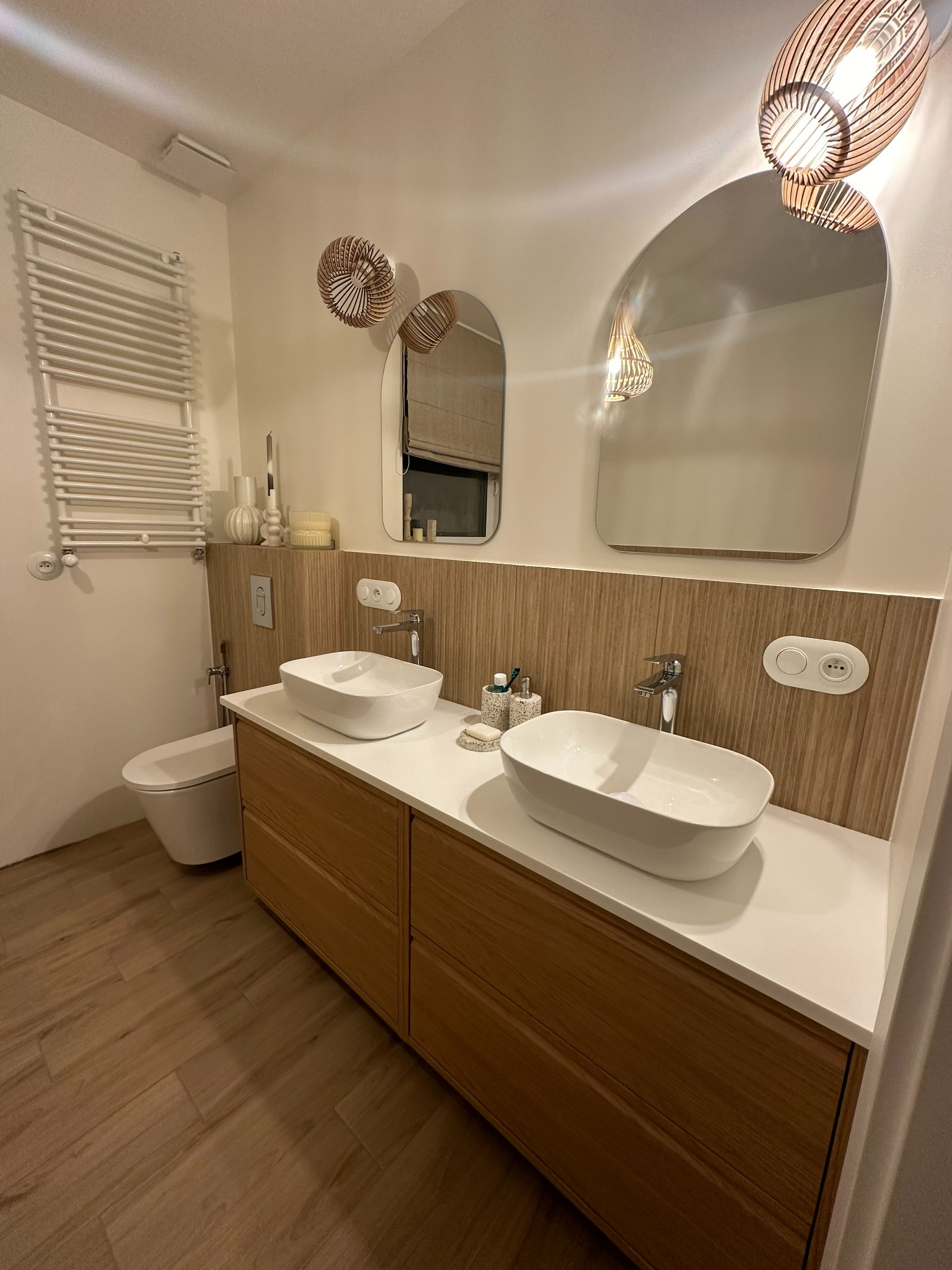 Nowoczesna łazienka z dwiema owalnymi umywalkami na drewnianej szafce, podłużne lustra i dekoracyjne lampy. Uwagę przykuwa drewniana boazeria i podłoga imitująca drewno.