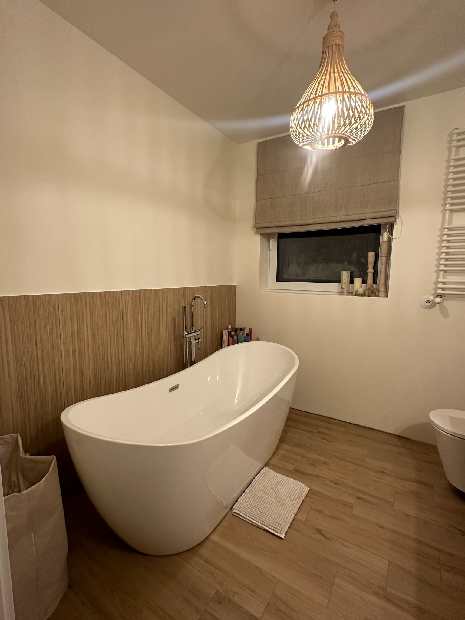 Nowoczesna łazienka z wolnostojącą wanną, drewnopodobnymi płytkami i dekoracyjną lampą wiszącą. Okno z roletą rzymską i świecami. Minimalistyczny grzejnik drabinkowy.