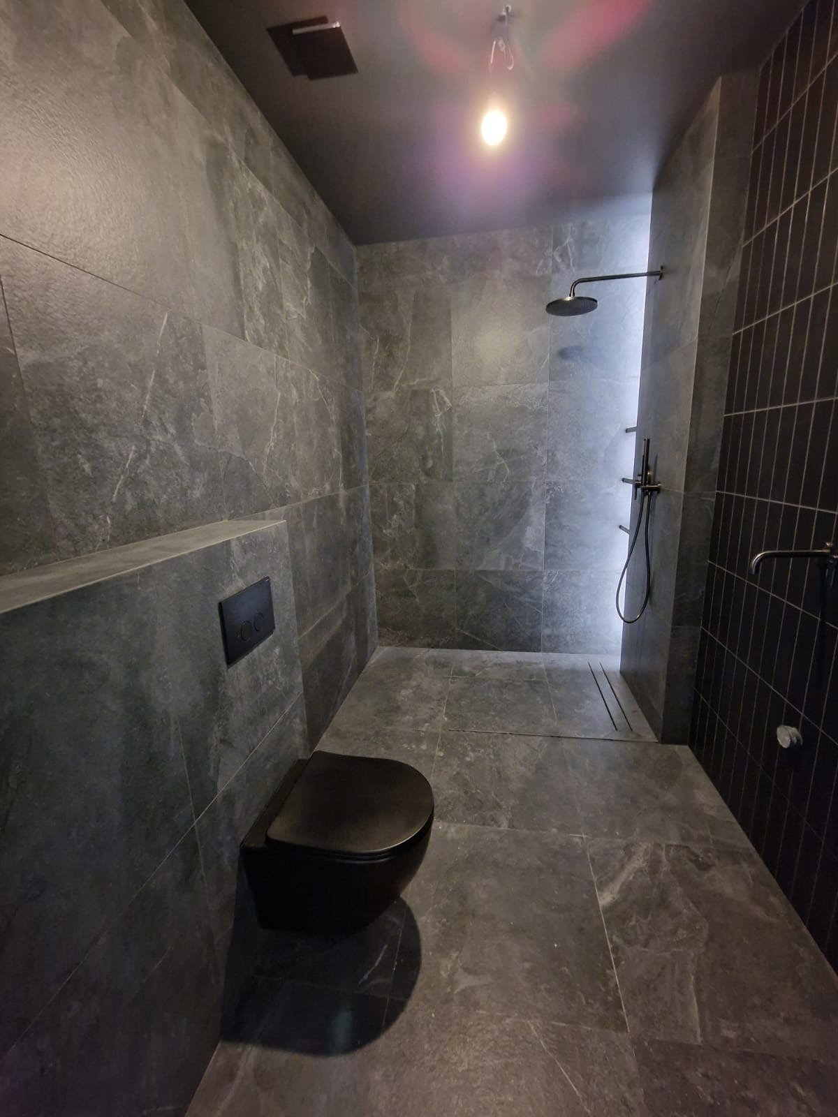 Nowoczesna łazienka w odcieniach szarości z czarną armaturą i podwieszaną toaletą. Minimalistyczny design, płytki imitujące kamień, oświetlenie punktowe.