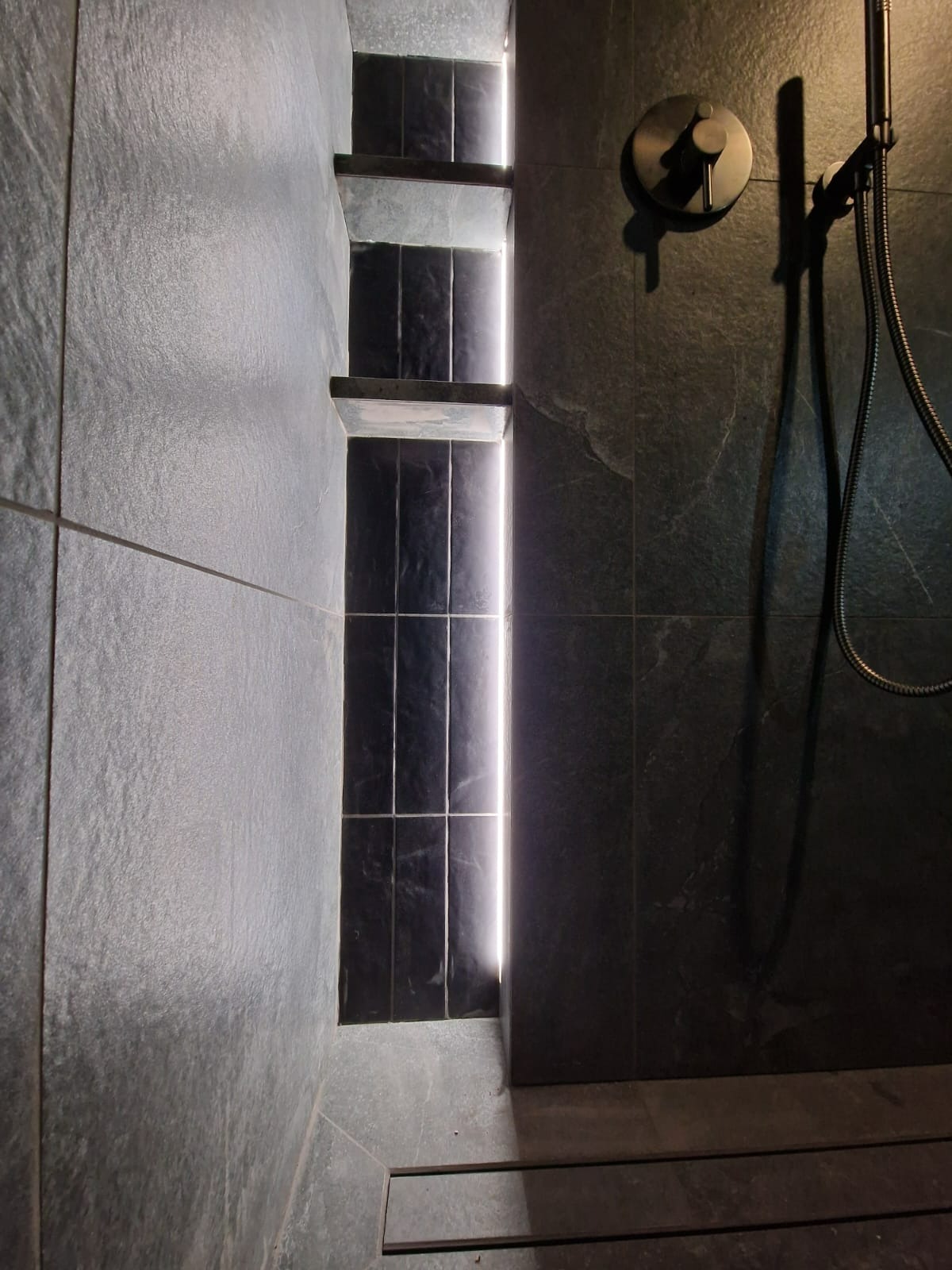 Nowoczesna kabina prysznicowa z czarnymi płytkami, wnęką z półkami podświetloną LED, deszczownicą i odpływem liniowym. Minimalistyczny design.