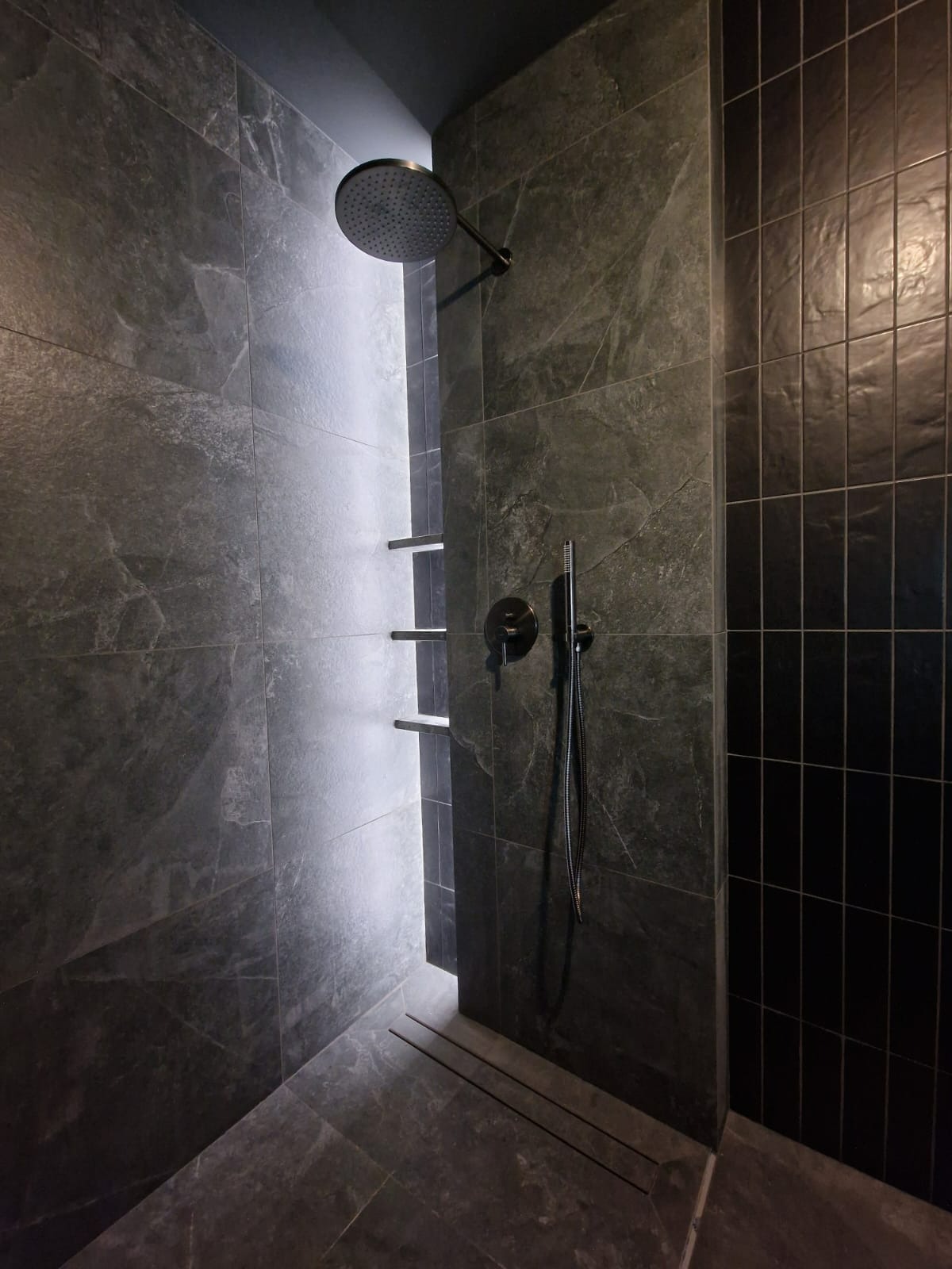 Nowoczesna kabina prysznicowa z szarymi płytkami imitującymi kamień, podświetlane półki w niszy i czarna armatura. Minimalistyczny design z odpływem liniowym.