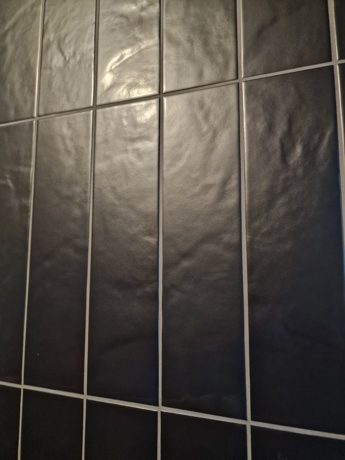 Ciemne, błyszczące, pionowo ułożone płytki ceramiczne z jasną fugą. Minimalistyczny wzór na ścianie. Widok pod kątem.