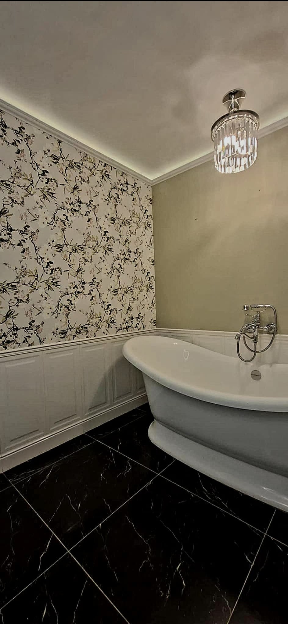 Elegancka łazienka z wolnostojącą wanną na czarnej marmurowej podłodze, ściana w kwiatowy wzór i kryształowy żyrandol. Klasyczny styl wykończenia wnętrza.