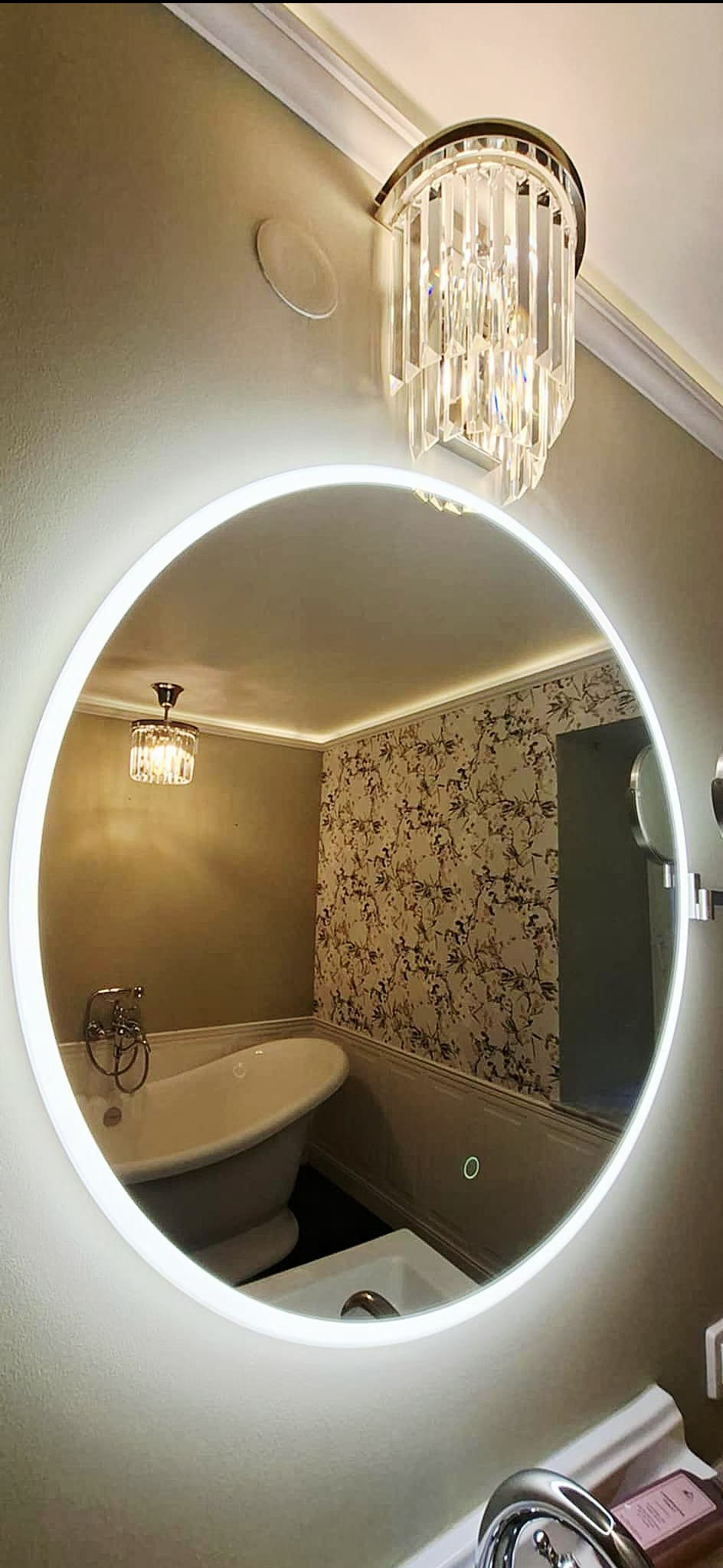 Okrągłe lustro LED odbija luksusową łazienkę z wolnostojącą wanną, wzorzystą tapetą i kryształowym żyrandolem. Nowoczesny design z nutą glamour.