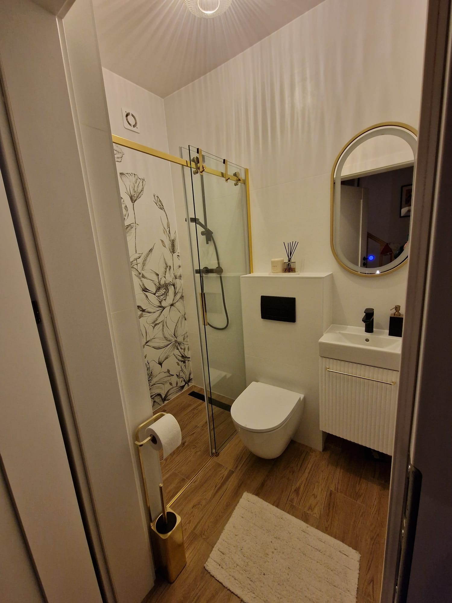 Nowoczesna łazienka z kabiną prysznicową ze złotymi detalami, umywalką z szafką i owalnym lustrem w złotej ramie. Na ścianie tapeta w kwiaty.