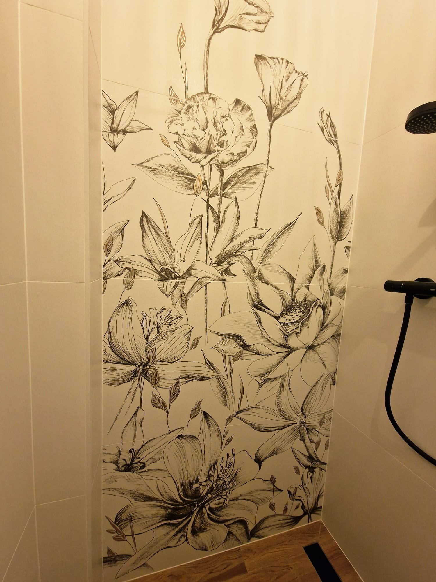 Beżowe płytki w łazience z motywem kwiatowym w stylu szkicu, czarna deszczownica i drewnopodobna podłoga z odpływem liniowym.