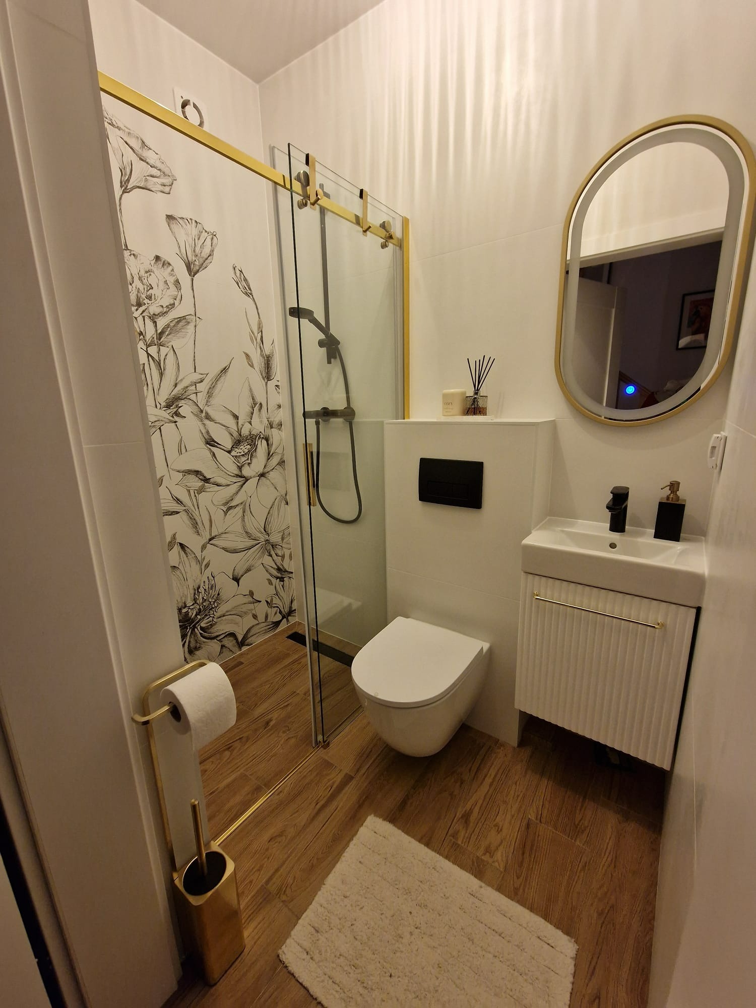 Nowoczesna łazienka z kabiną prysznicową ze złotymi akcentami, umywalką z szafką, toaletą i drewnopodobną podłogą. Na ścianie lustro w złotej ramie.
