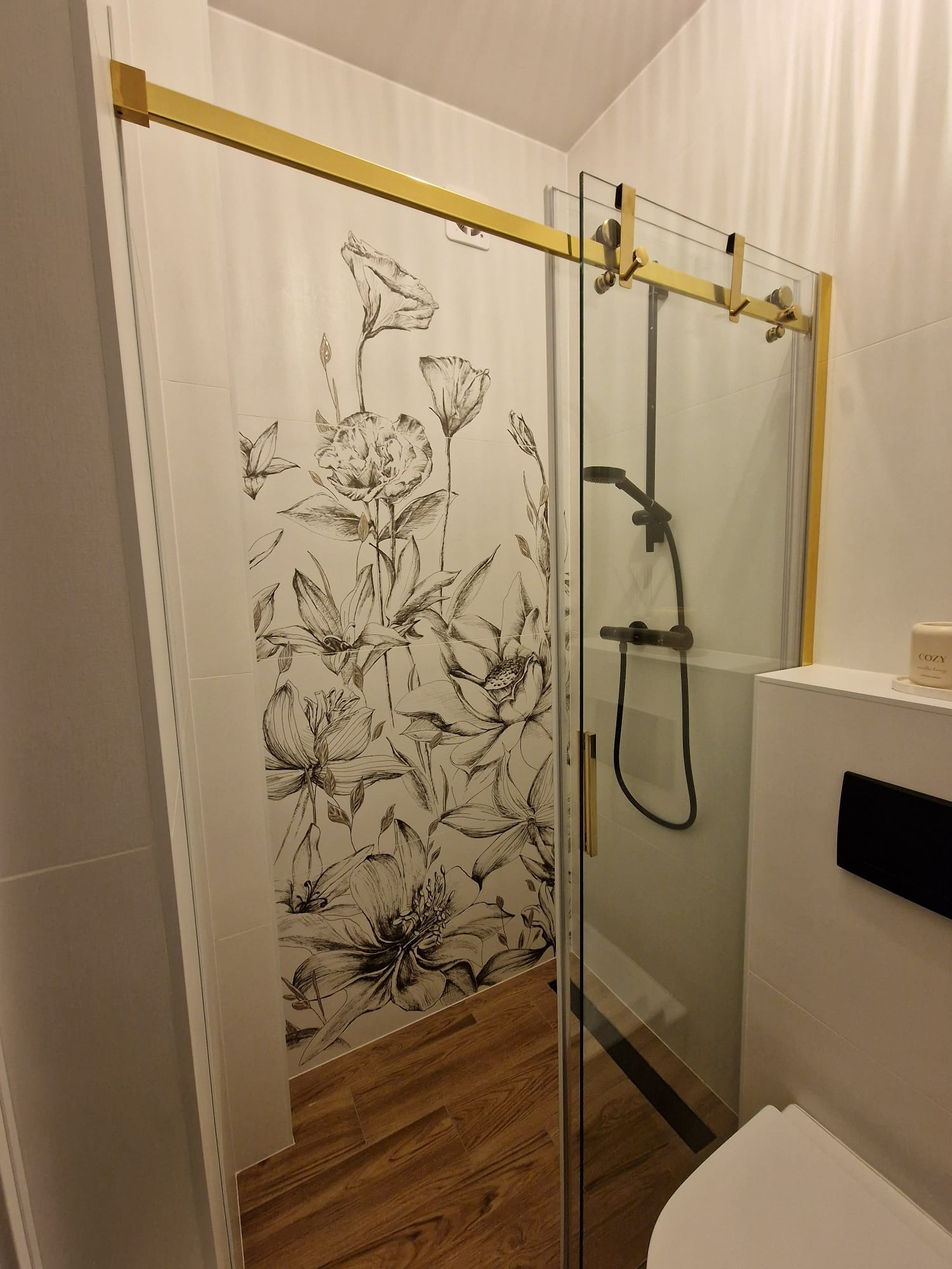Nowoczesna kabina prysznicowa z przesuwnymi drzwiami, złote detale, płytki z motywem kwiatowym, drewnopodobna podłoga, minimalistyczny design.