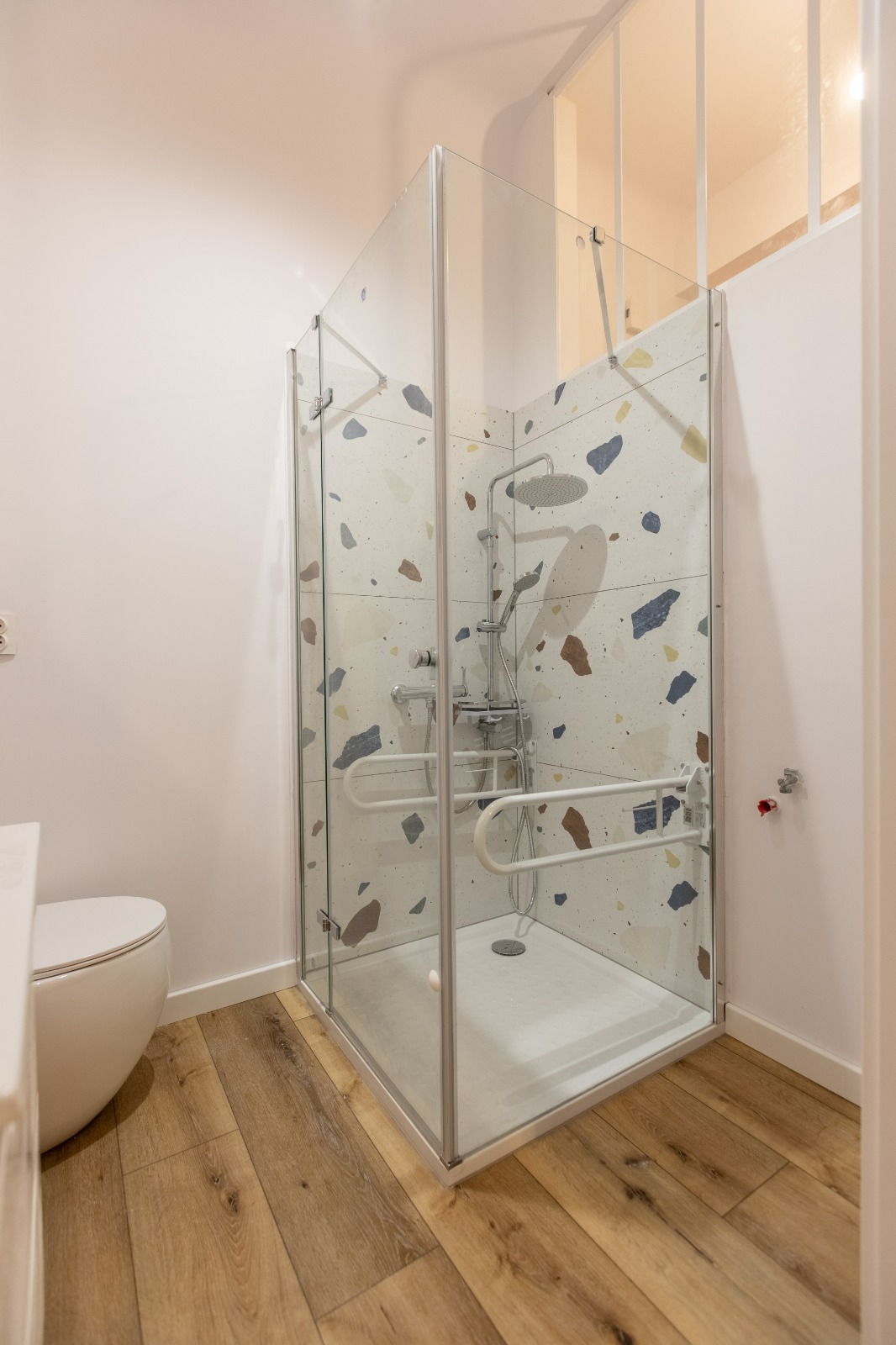 Nowoczesna kabina prysznicowa z uchwytami dla niepełnosprawnych, wykończona płytkami w stylu terrazzo, na drewnopodobnej podłodze. Jasne, minimalistyczne wnętrze łazienki.