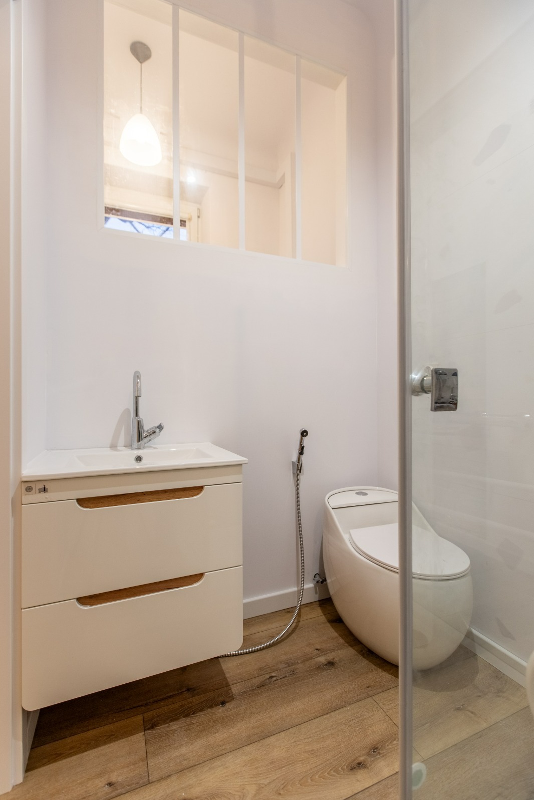 Nowoczesna, jasna łazienka z drewnianą podłogą. Umywalka z szafką, toaleta z bidetem i kabina prysznicowa. Okno z luksferami w górnej części ściany.