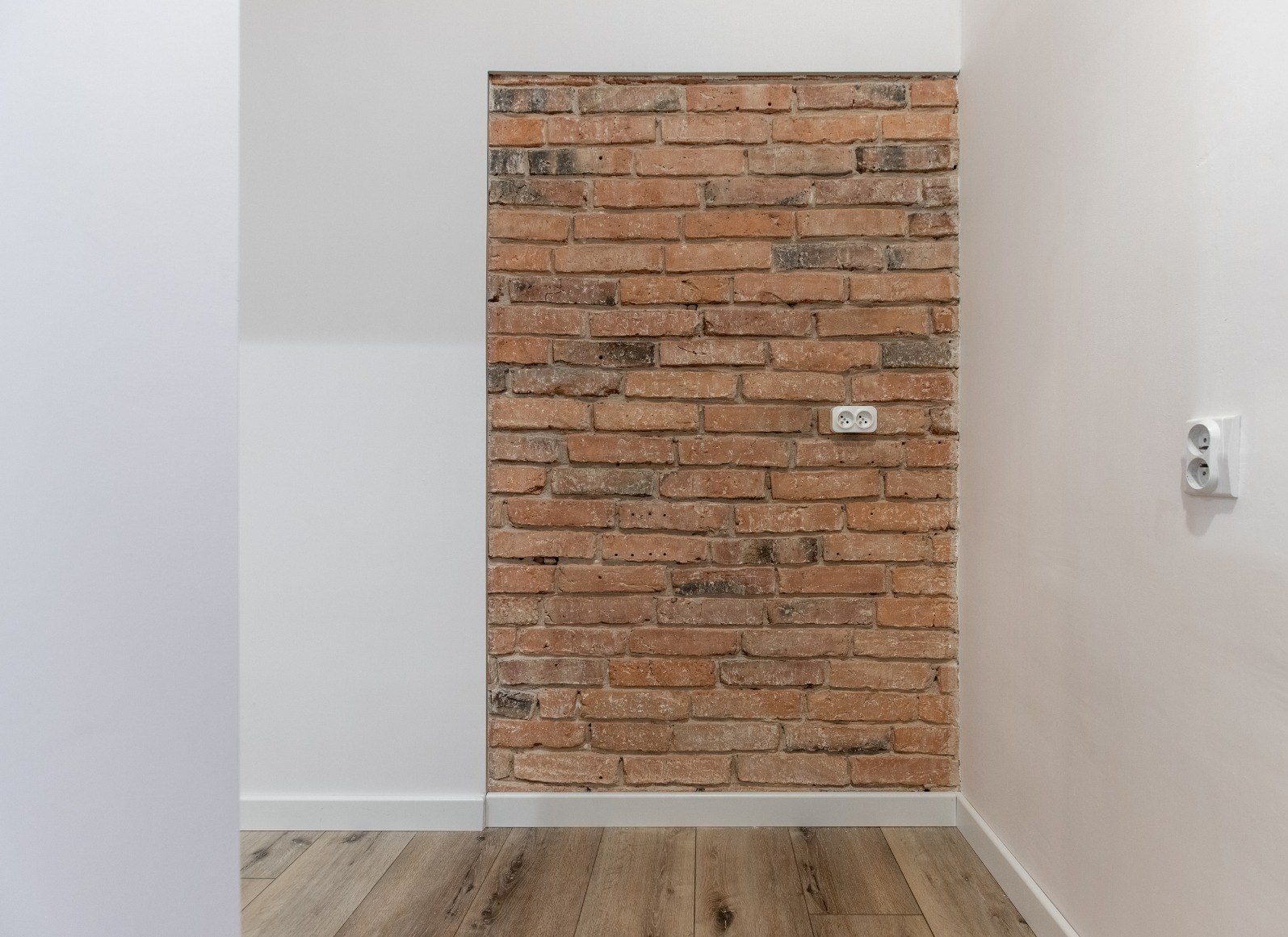 Ceglana ściana z gniazdkami w odnowionym wnętrzu. Widoczne drewniane panele podłogowe i białe ściany. Akcent na połączenie surowego muru z nowoczesnym wykończeniem.