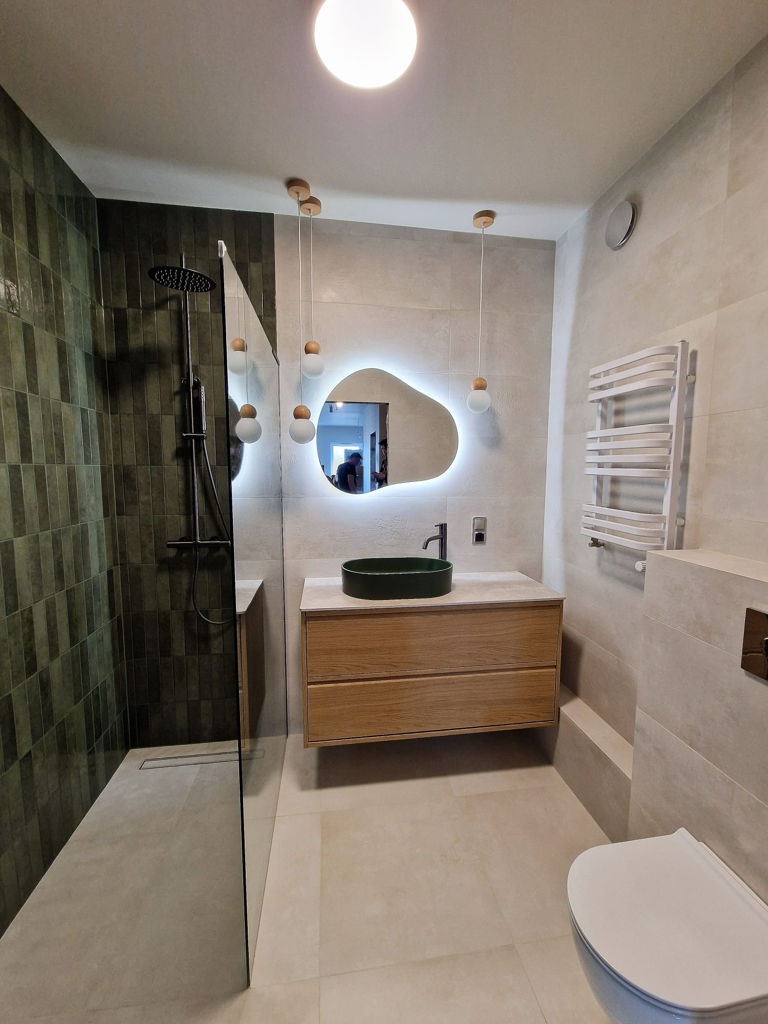 Nowoczesna łazienka z kabiną prysznicową typu walk-in, umywalką w kolorze butelkowej zieleni i podświetlanym lustrem o nieregularnym kształcie. Minimalistyczny design.