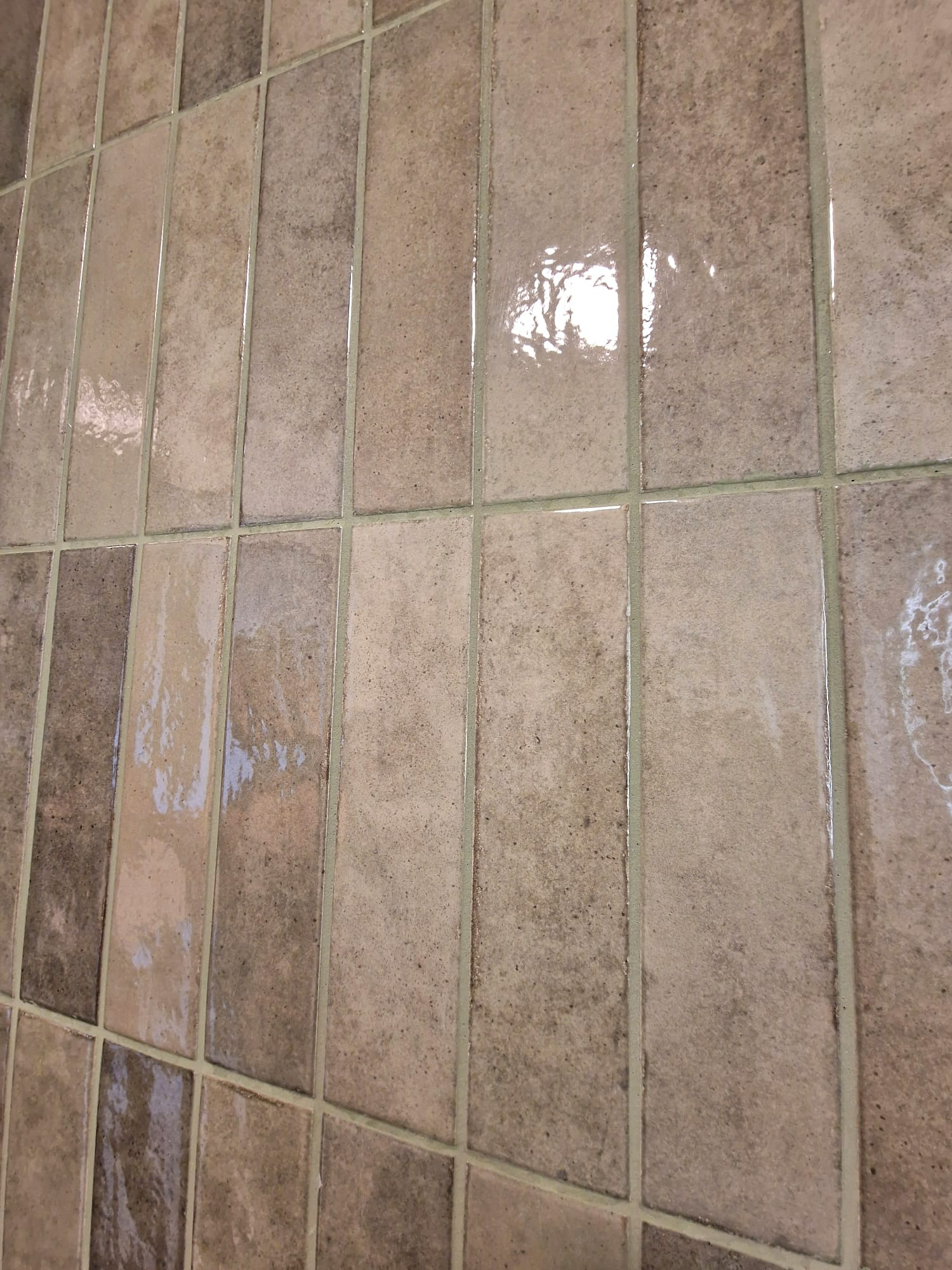 Błyszczące, beżowo-brązowe, wąskie płytki ułożone pionowo na ścianie. Widoczne fugi w kolorze zbliżonym do płytek. Odbicie światła na powierzchni płytek.