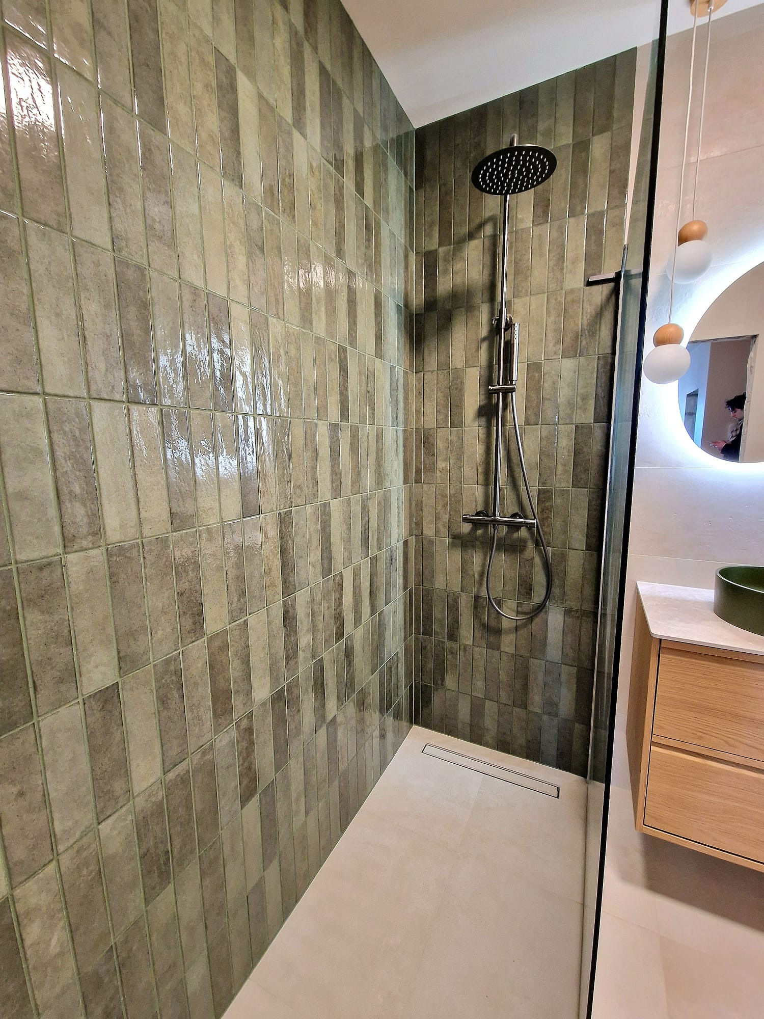 Nowoczesna kabina prysznicowa z deszczownicą i odpływem liniowym, wykończona podłużnymi, błyszczącymi płytkami w odcieniach szarości i beżu. Umywalka z drewnianą szafką.