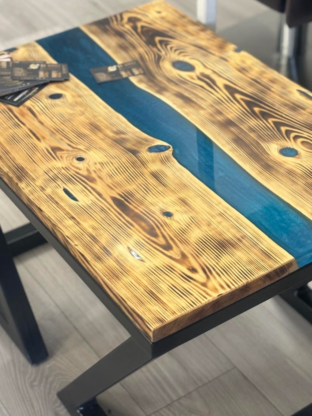 Stolik kawowy z blatem z wypalanego drewna z żywicą epoksydową w kolorze turkusowym, czarne metalowe nogi, wizytówki ułożone na blacie.