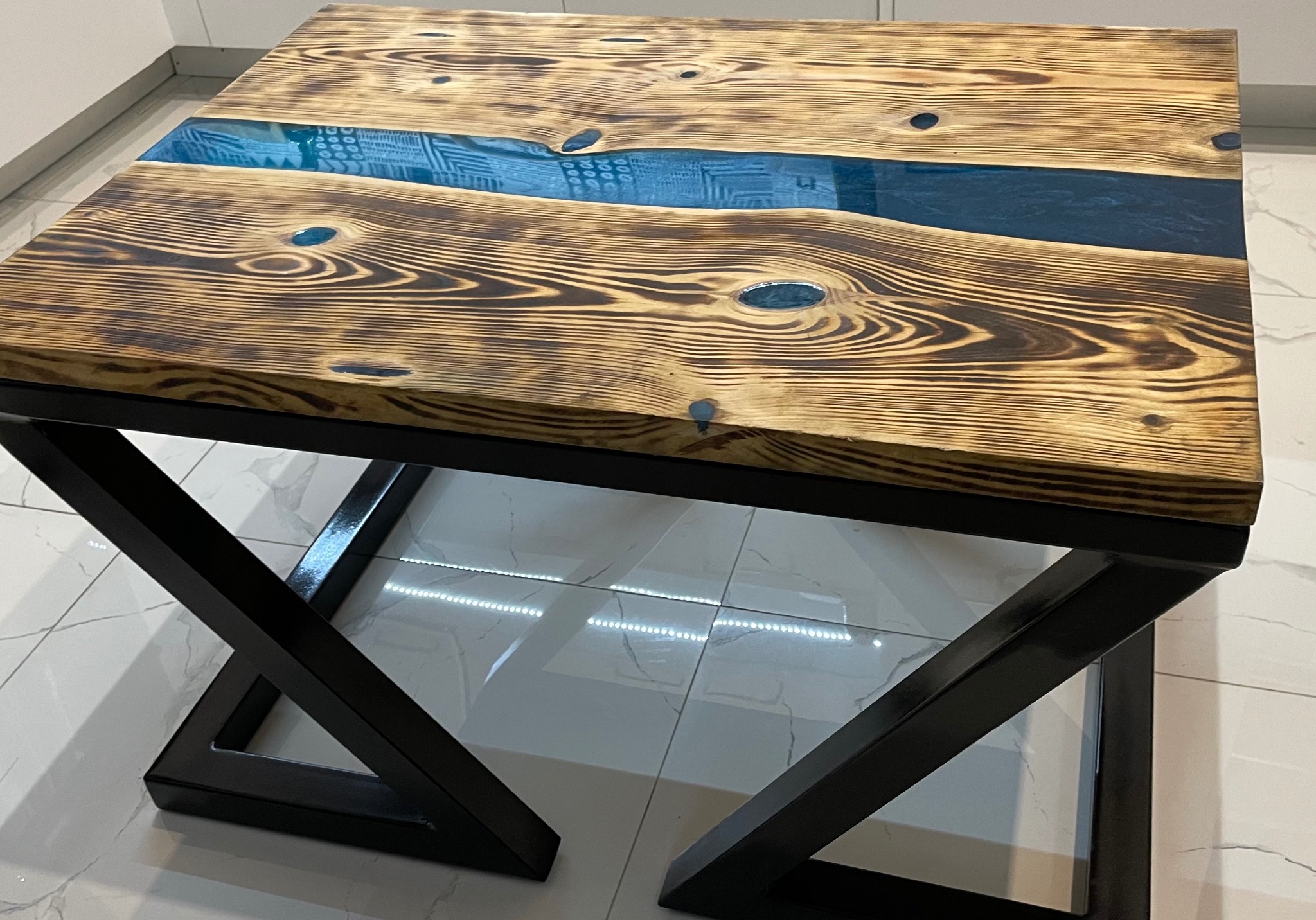 Stolik kawowy z blatem z palonego drewna z żywicą epoksydową w kolorze turkusowym, osadzony na czarnych, metalowych nogach w kształcie litery X, na tle białych płytek podłogowych.