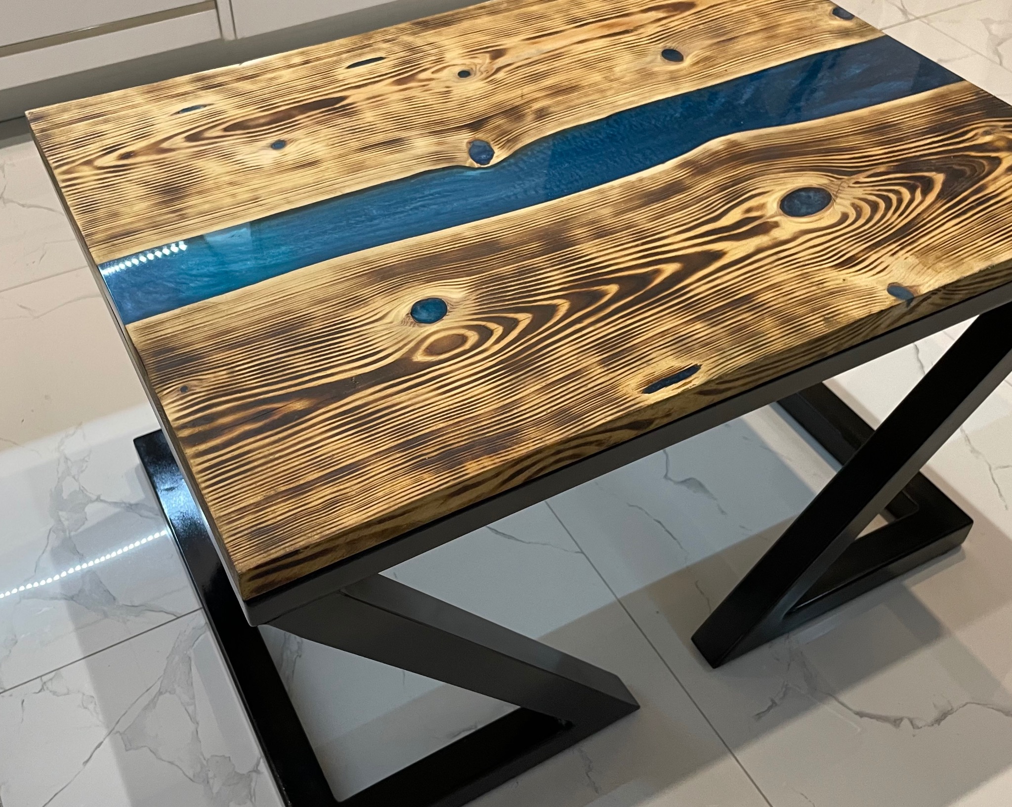 Stół z blatem z wypalanej deski z żywicą epoksydową w kolorze turkusowym, czarne metalowe nogi o geometrycznym kształcie, widok z góry na marmurowej podłodze.