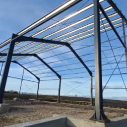 Stalowa konstrukcja hali w trakcie budowy, widok z dołu na szkielet z belek i słupów na betonowym fundamencie, błękitne niebo w tle.