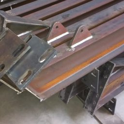 BEDSTALL Marlena Biedra - Stalowe elementy konstrukcyjne, profile i blachy z otworami, ułożone warstwami na podłodze. Widoczne ślady spawania i obróbki metalu.