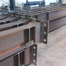 BEDSTALL Marlena Biedra - Stalowe belki konstrukcyjne z przyspawanymi elementami, przygotowane do montażu. Widoczne otwory na śruby. W tle maszyny do obróbki metalu.