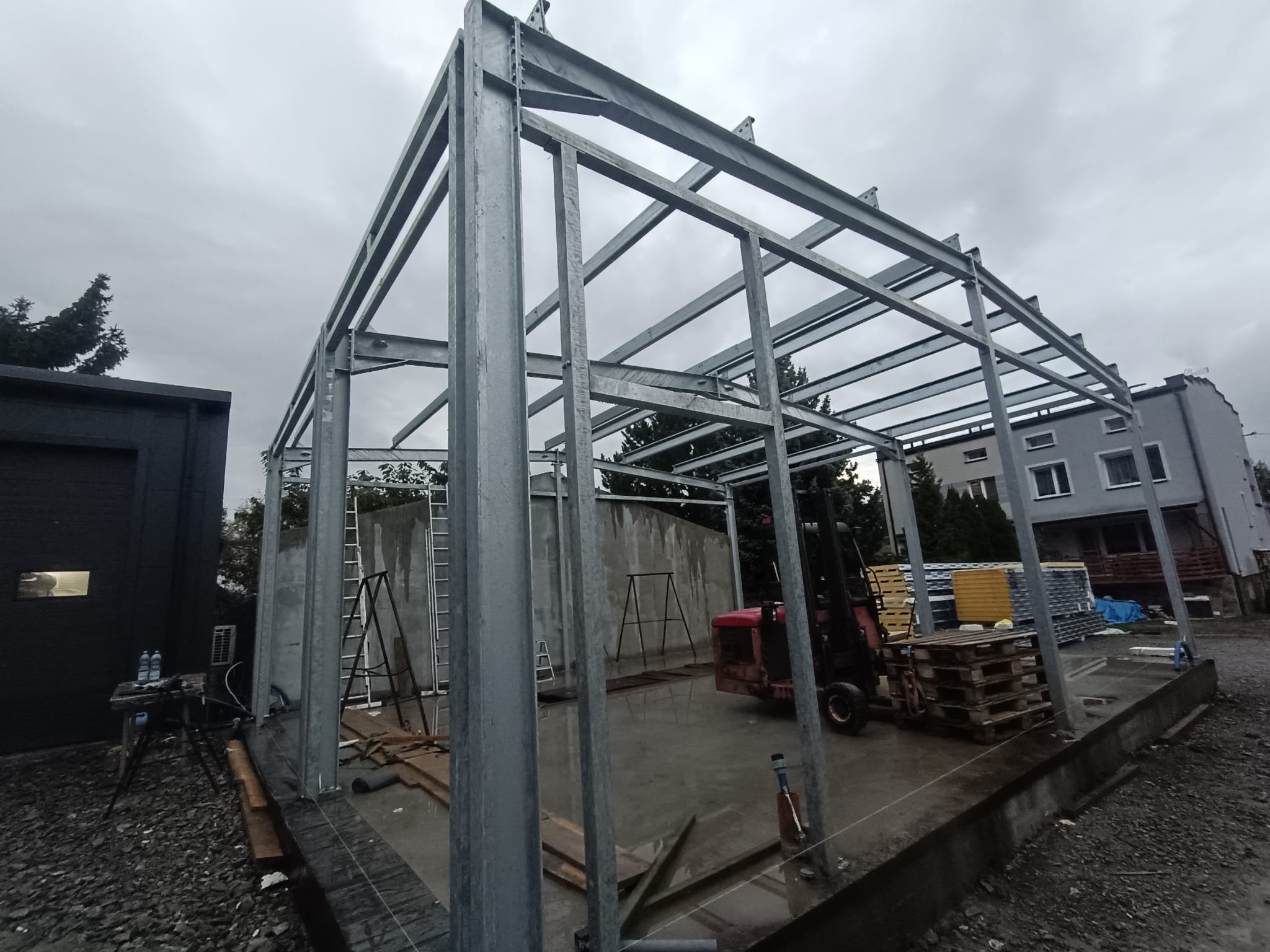 Konstrukcja stalowa hali w trakcie budowy, widoczne elementy nośne, słupy i dźwigary, na zewnątrz, pochmurny dzień, w tle budynki i drzewa.