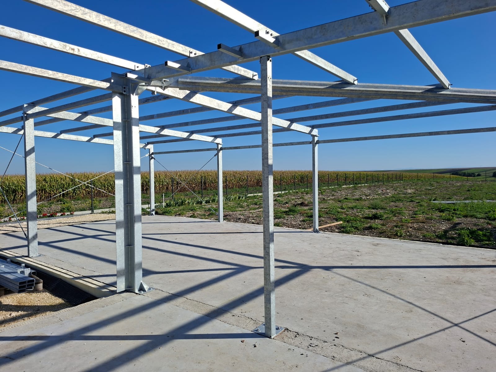 Metalowa konstrukcja hali na betonowej płycie, widok na pole kukurydzy pod błękitnym niebem. Elementy konstrukcji skręcane śrubami.