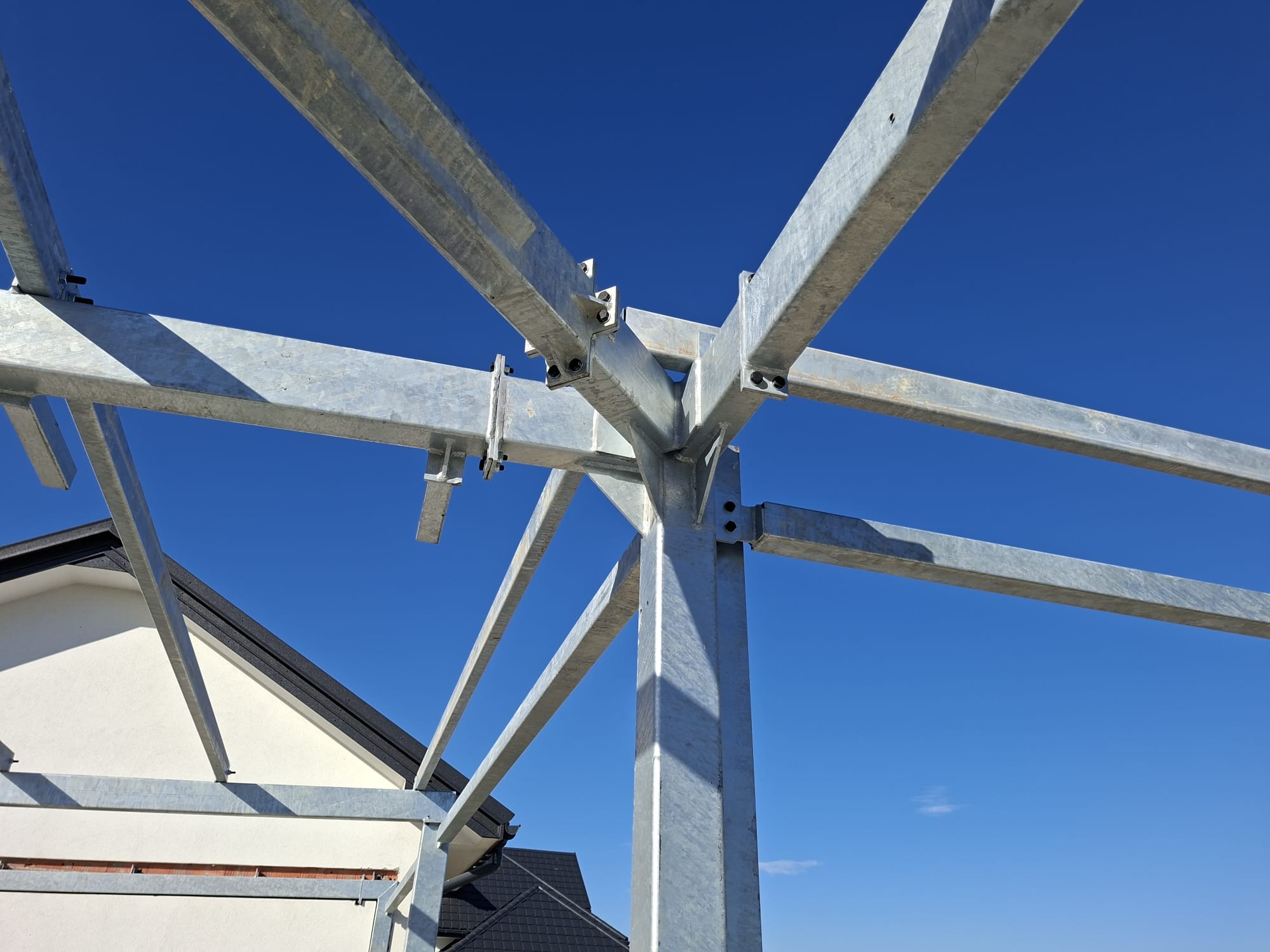 Konstrukcja stalowa z profili ocynkowanych, skręcana śrubami, na tle błękitnego nieba i fragmentu budynku. Widok z dołu, podkreślający geometrię połączeń.