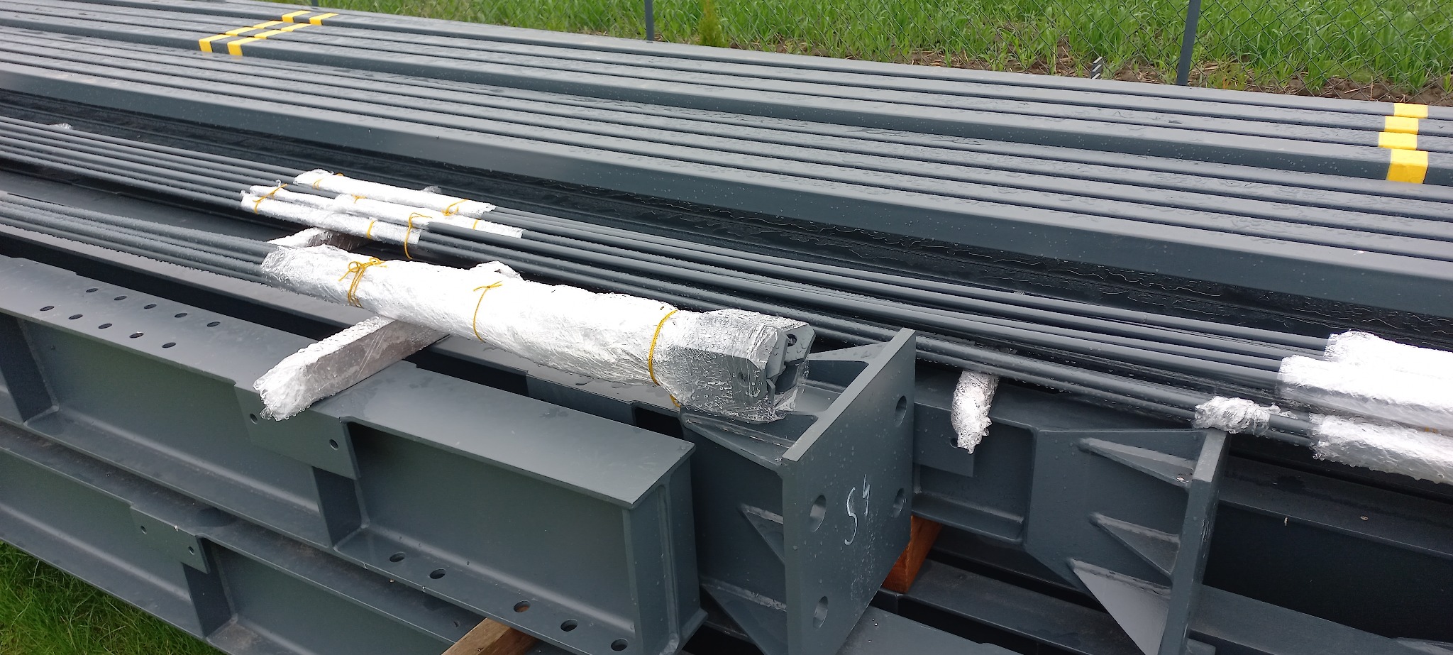 Stalowe elementy konstrukcyjne, belki i pręty, przygotowane do budowy hali, zabezpieczone folią i taśmą, ułożone na trawie. Widoczne ślady wilgoci.