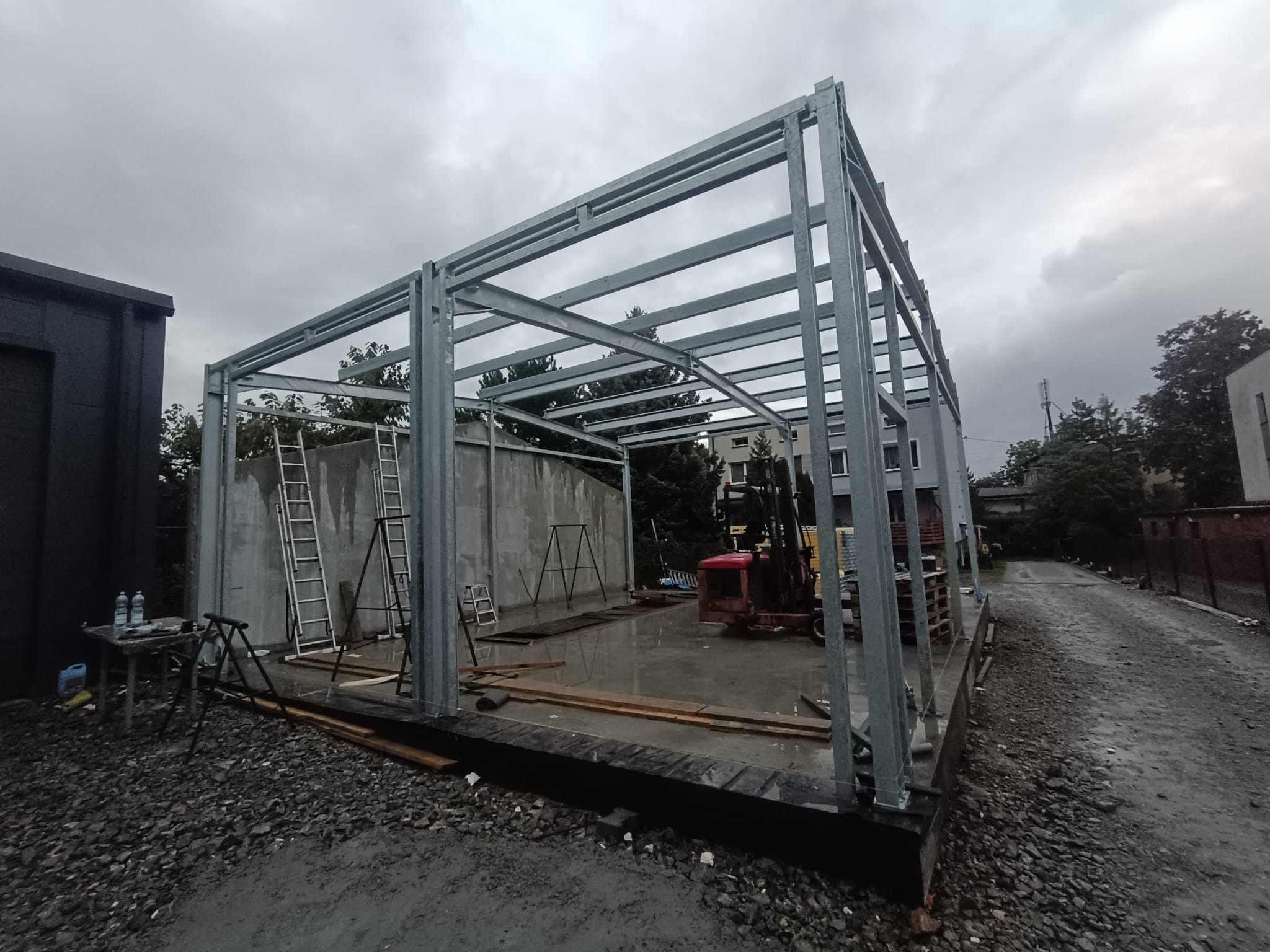 Konstrukcja stalowa hali w trakcie budowy, widoczne drabiny, sprzęt budowlany i betonowa ściana w tle, pochmurne niebo.