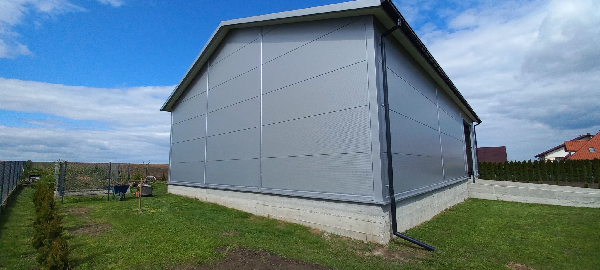 Szara hala z paneli warstwowych, widok z zewnątrz na tle błękitnego nieba. Widoczne detale konstrukcyjne i otoczenie hali: trawnik, drzewa, inne budynki w tle.