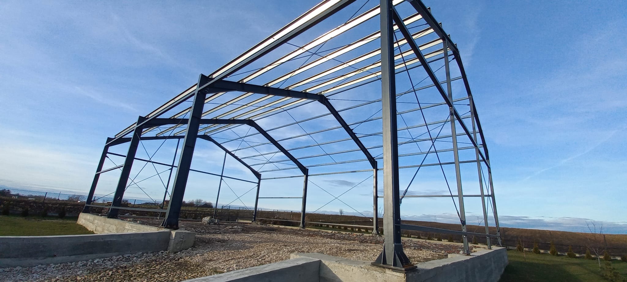 Stalowa konstrukcja hali w trakcie budowy, widok z dołu na szkielet z belek i słupów na betonowym fundamencie, błękitne niebo w tle.