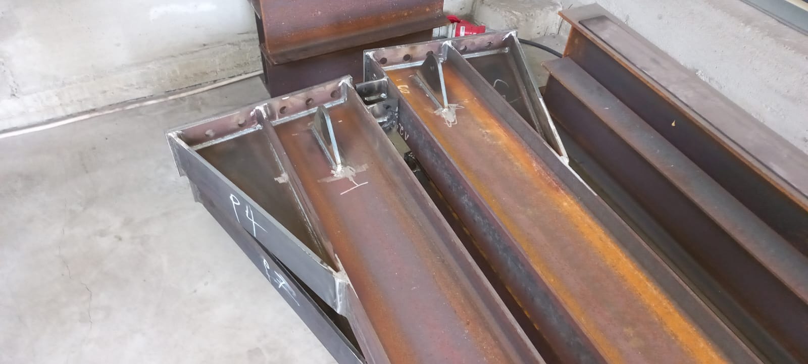 Metalowe elementy konstrukcyjne z widocznymi spawami i otworami, przygotowane do dalszego montażu. Widoczne oznaczenia 'P4' i 'REV' na jednym z elementów.