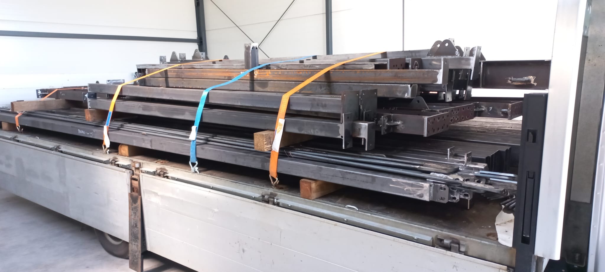 Stalowe elementy konstrukcyjne, zabezpieczone pasami transportowymi, na platformie pojazdu dostawczego. Widoczne spawy i otwory montażowe.