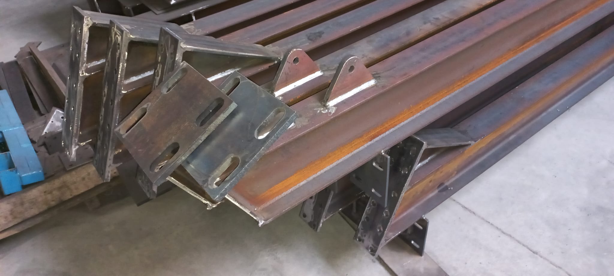 Stalowe elementy konstrukcyjne, profile i blachy z otworami, ułożone warstwami na podłodze. Widoczne ślady spawania i obróbki metalu.