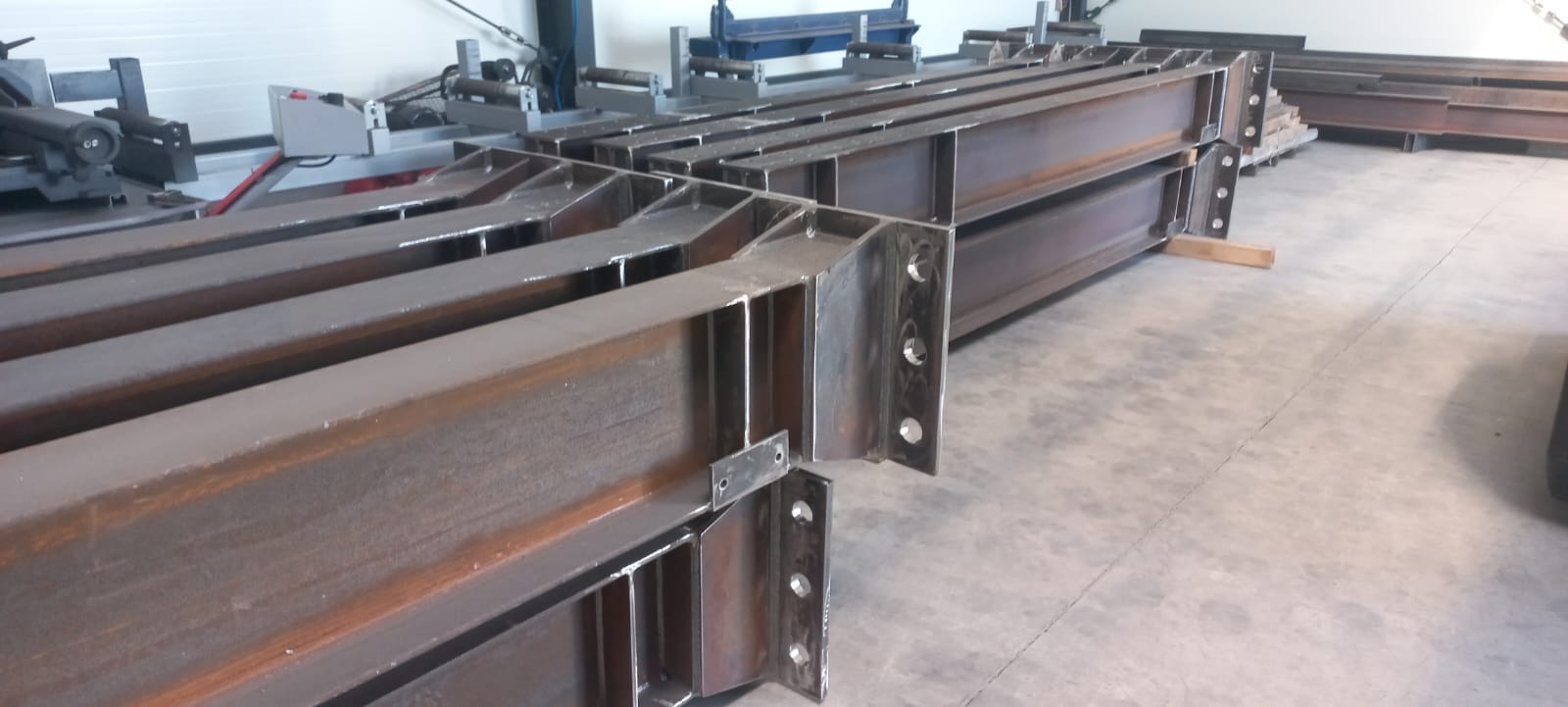 Stalowe belki konstrukcyjne z przyspawanymi elementami, przygotowane do montażu. Widoczne otwory na śruby. W tle maszyny do obróbki metalu.